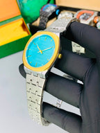 A wristwatch with a bright blue dial, gold-tone bezel and hands, and a silver-tone metal link bracelet, held by a hand wearing a black glove. The background shows a light gray surface with other watches and colorful boxes blurred behind it. Kustore