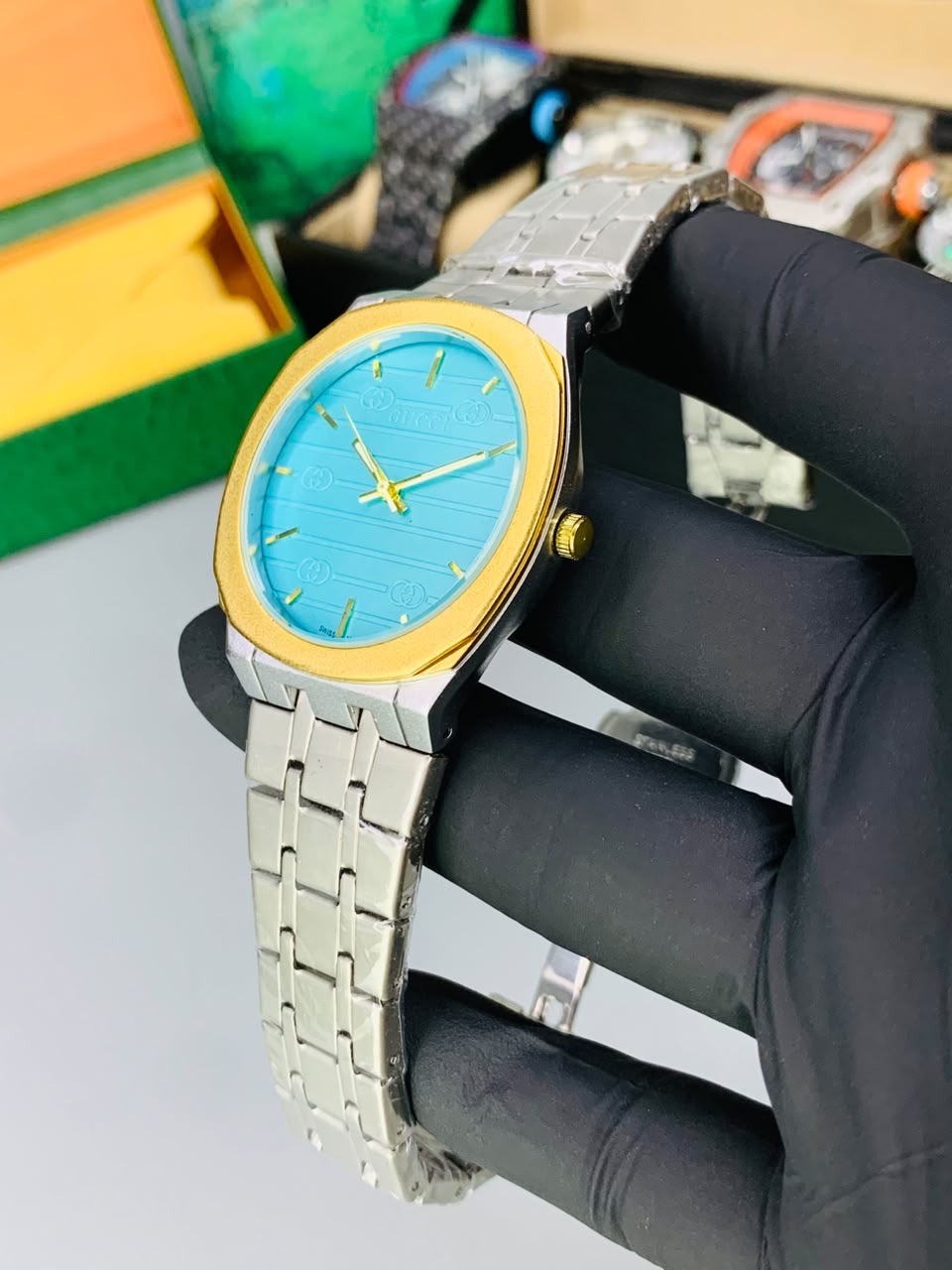 A wristwatch with a bright blue dial, gold-tone bezel and hands, and a silver-tone metal link bracelet, held by a hand wearing a black glove. The background shows a light gray surface with other watches and colorful boxes blurred behind it. Kustore