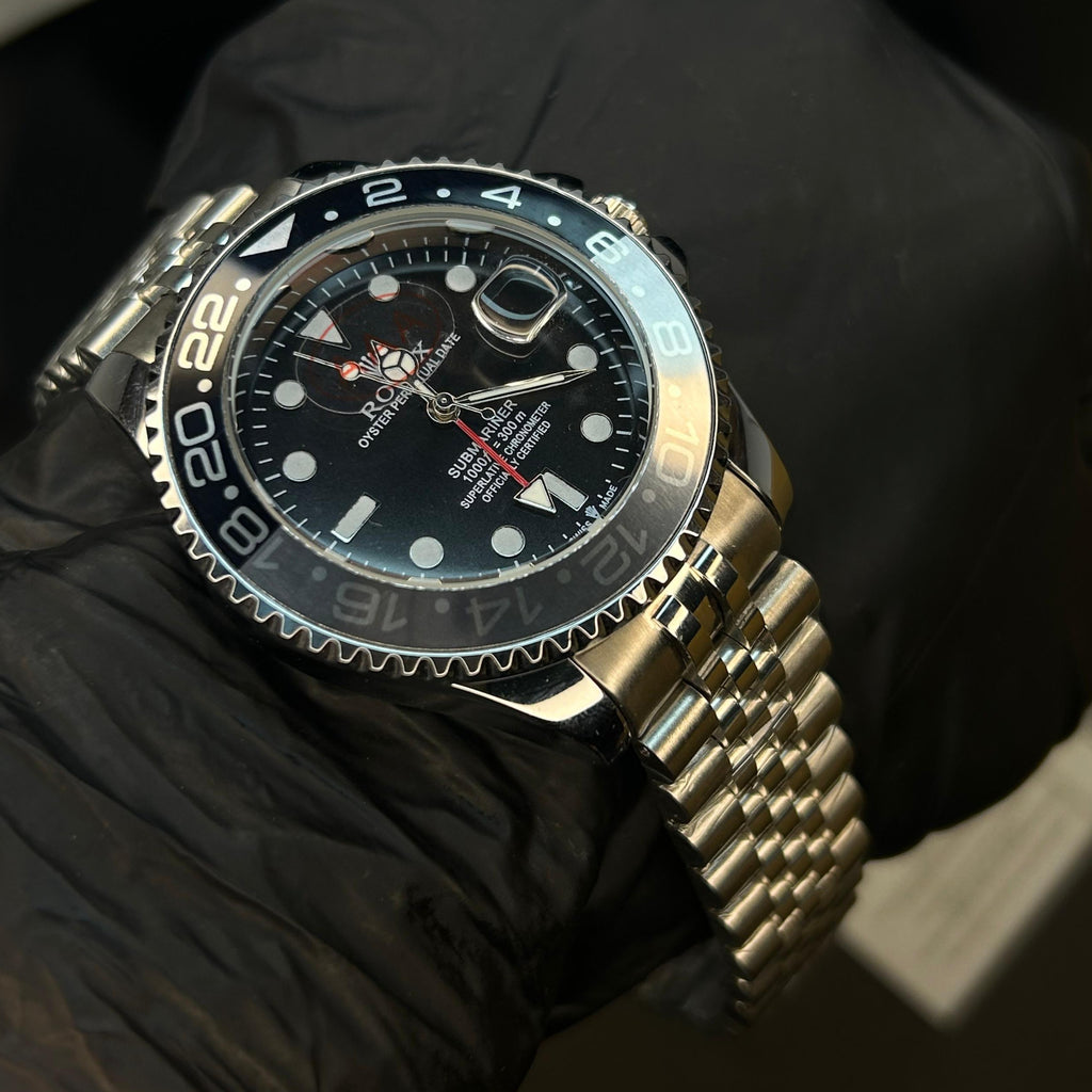 A stainless steel wristwatch with a black dial and bezel, worn on a hand covered by a black glove. The watch features a metal link bracelet and a black bezel with white numerals.