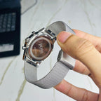 A close-up of a hand holding the back of a silver BOSS watch with a stainless steel mesh band, showing the branded case end and clasp, against a light marble surface.